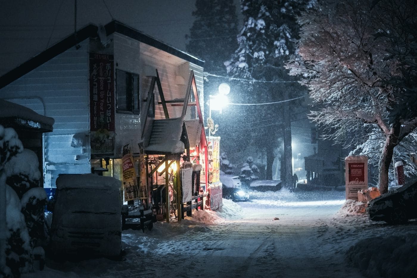 A quiet side street in Hakuba at night, with snow falling, snowbanks and lit shopfronts