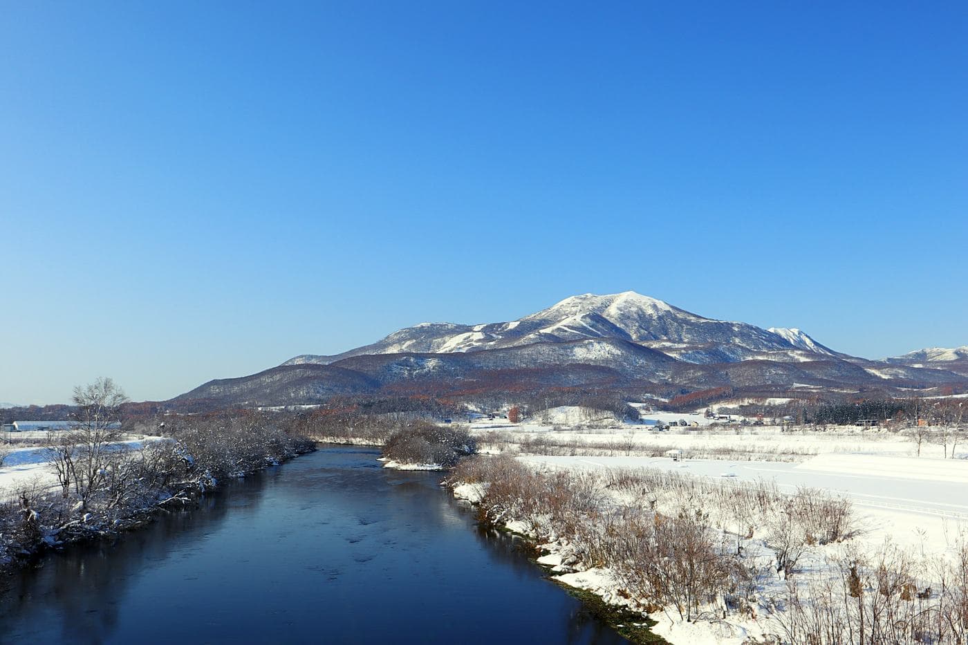 Snow-covered Niseko Annupuri behind a river and white fields, seen from Kutchan on a clear day